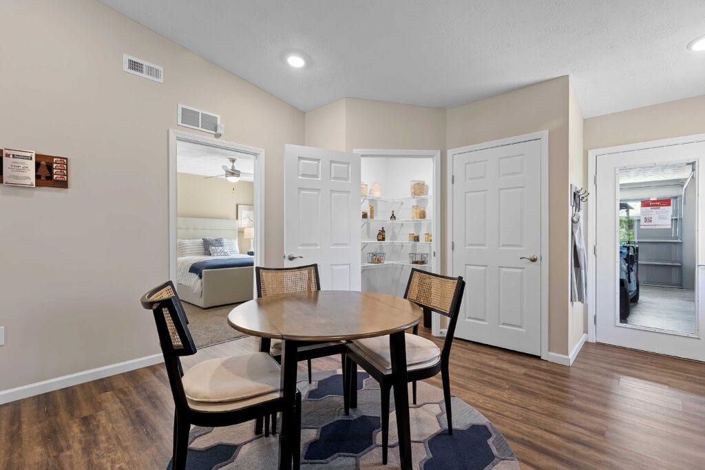 A round wooden dining table with four chairs on a rug in a room with wood flooring, beige walls, and open doors leading to a bedroom, pantry, and a sunroom.