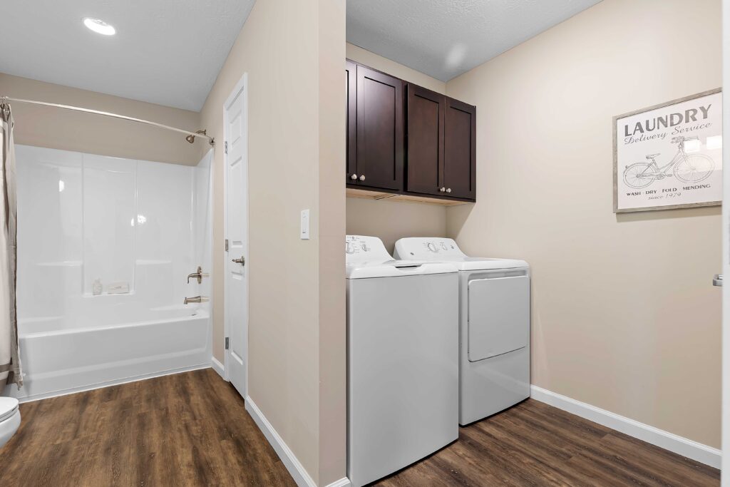 A laundry room with a washer and dryer next to a bathroom with a bathtub and shower, wood-style flooring, beige walls, and dark wood cabinets above the machines.