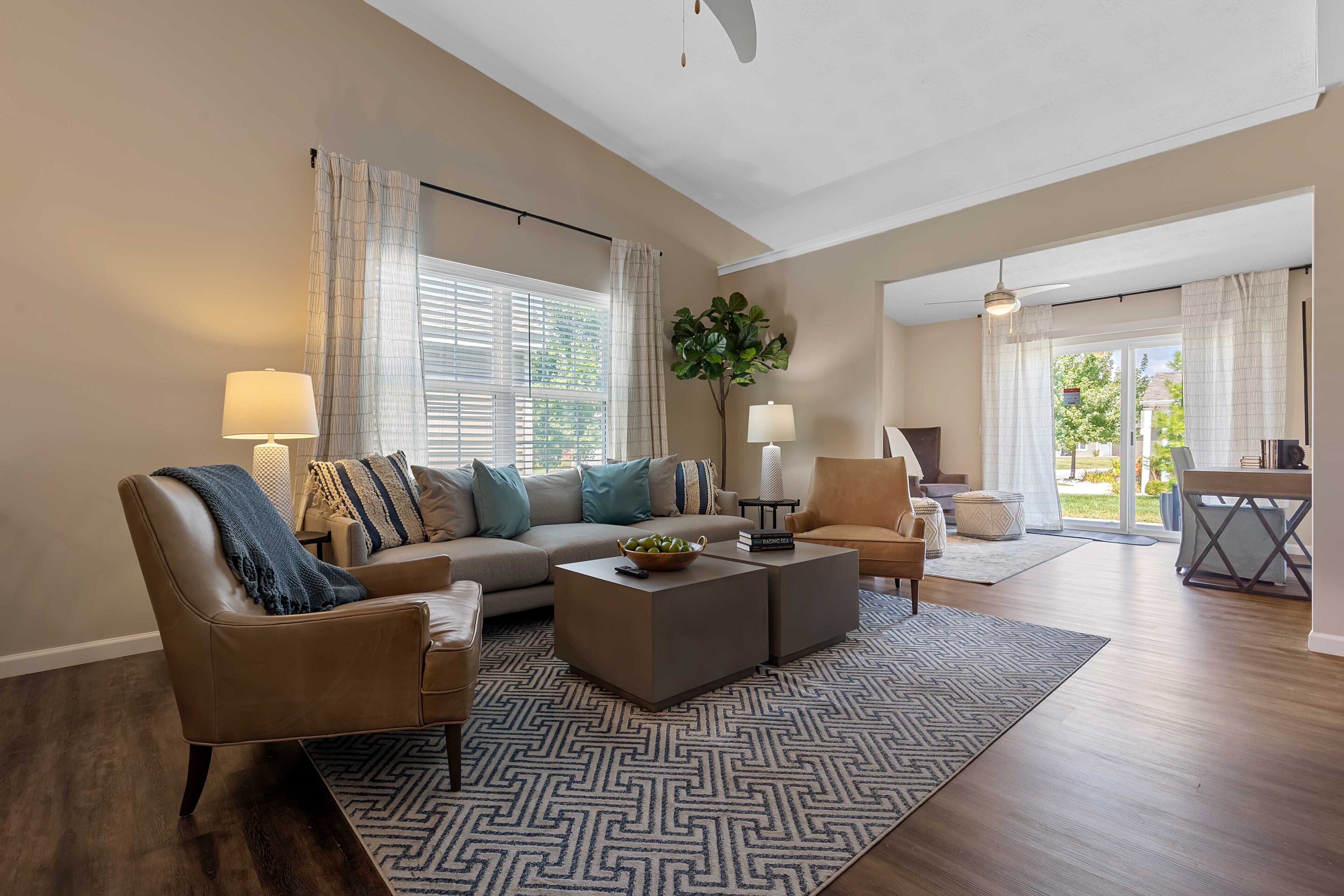 Living room with a gray sectional sofa, leather armchair, patterned rug, two lamps, large windows with curtains, and a view into a bright adjacent room.