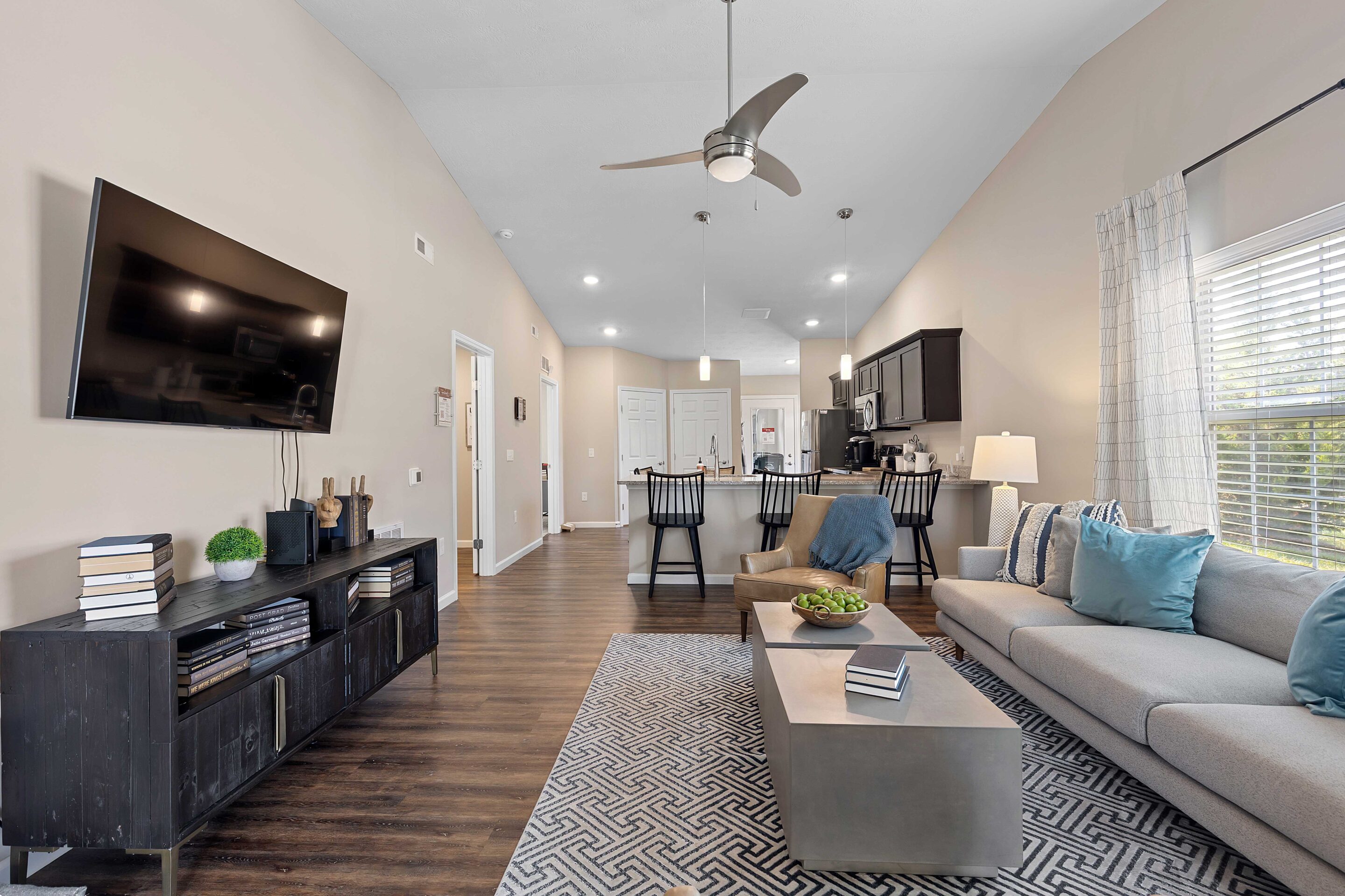 Modern open-concept living room and kitchen with a mounted TV, gray sectional sofa, black cabinets, barstools, and large windows letting in natural light.