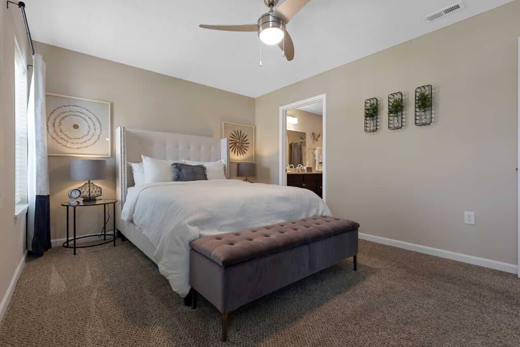 A modern bedroom with a white bed, gray upholstered bench, two side tables with lamps, wall art, decor plants, and an attached bathroom visible through an open door.