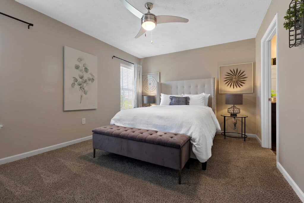A neatly made bed with white linens sits in a carpeted bedroom featuring beige walls, modern artwork, a gray tufted bench, and a ceiling fan with light.