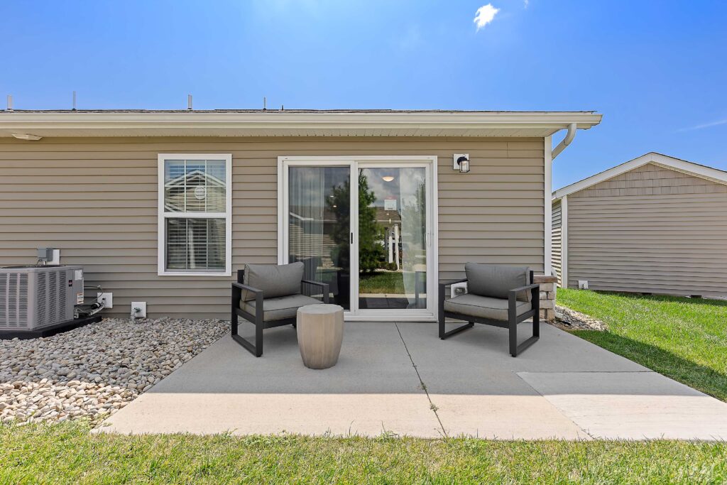 A small concrete patio with two chairs and a round table sits outside a house with beige siding and a sliding glass door. Grass and rocks border the patio.
