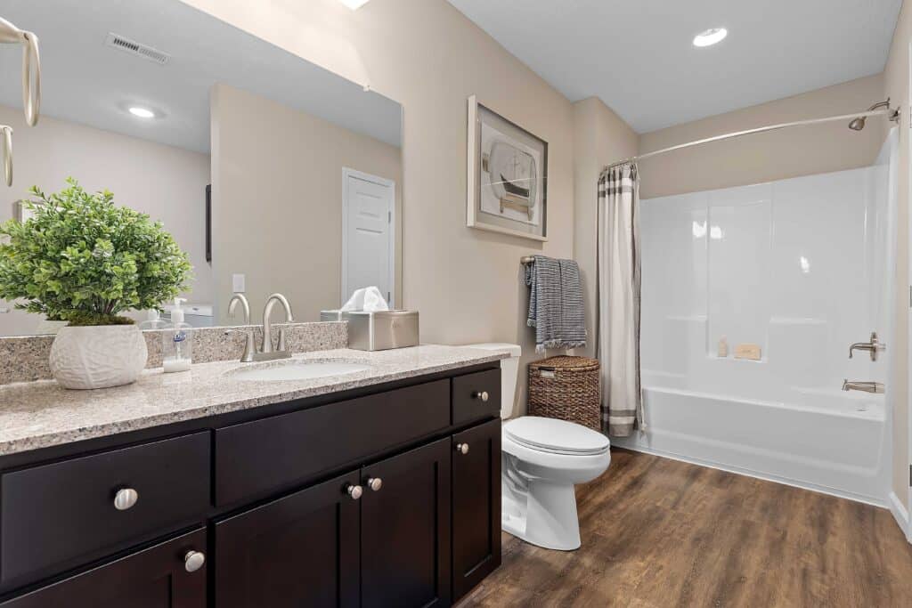Modern bathroom with dark wood vanity, granite countertop, toilet, white bathtub-shower combo, potted plant, and wicker basket on wood flooring.