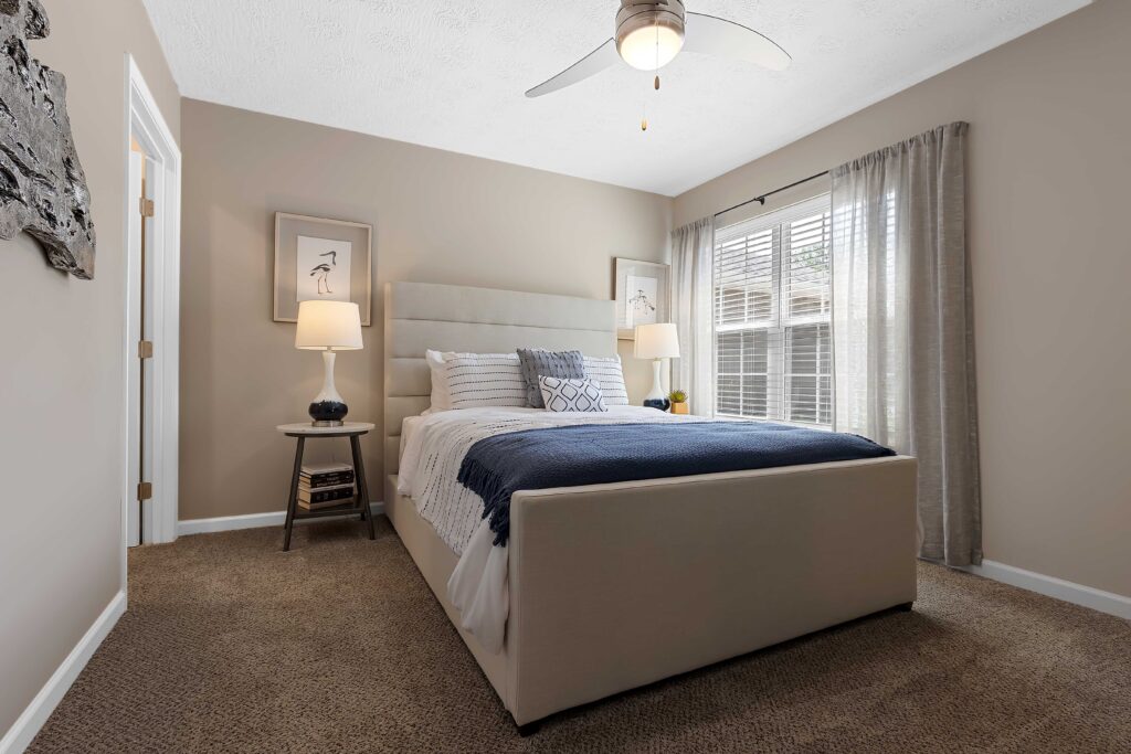 A neatly made bed with blue and white bedding is centered in a modern bedroom with beige walls, carpet, two nightstands, lamps, and a ceiling fan. Light enters through a window with curtains.