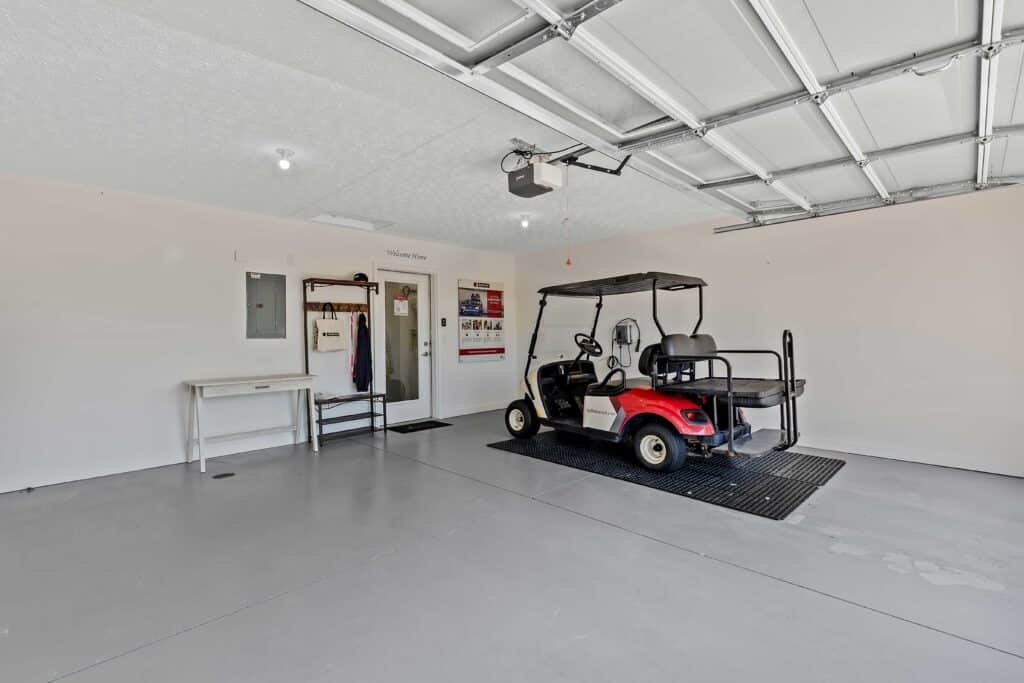 A clean, spacious garage with a red and black golf cart parked inside, a small table, a coat rack with hanging jackets, and utility panels on the wall.