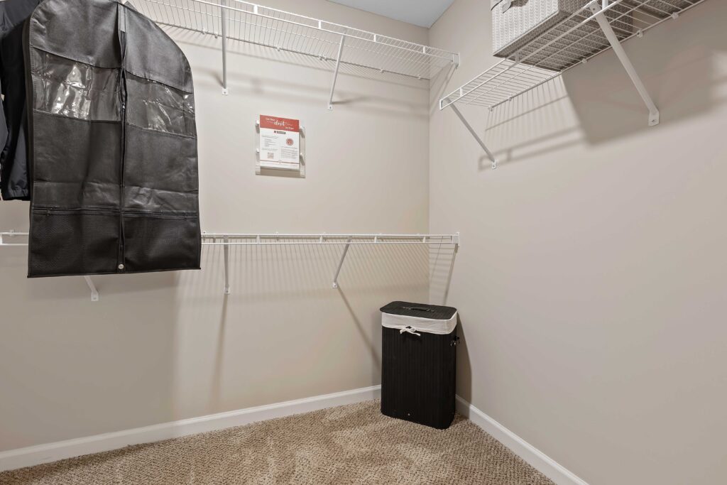 Walk-in closet with beige carpet, empty white wire shelves, a black laundry hamper, and a suit bag hanging on the left.