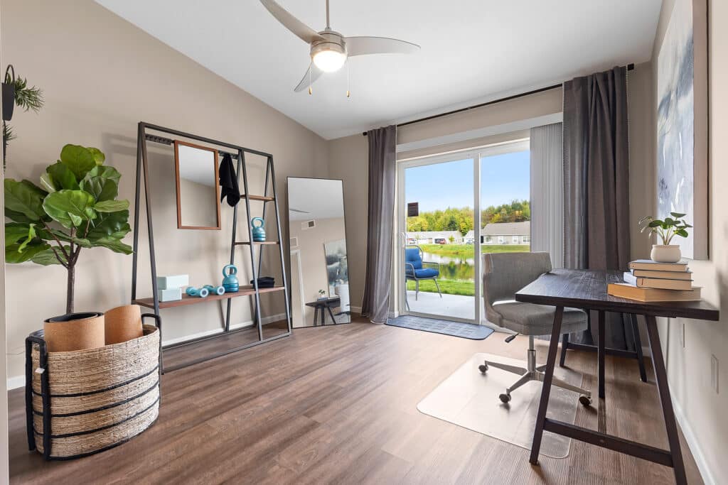 A home office with a desk, chair, and books, a large mirror on a stand, a basket with yoga mats, a potted plant, and sliding glass doors opening to a balcony with a grassy view.