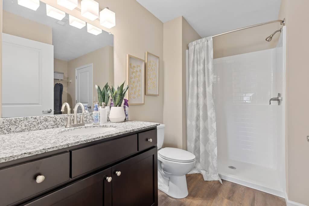 Modern bathroom with a granite countertop, dark wood vanity, toilet, walk-in shower with white tile, and decorative plants and artwork.