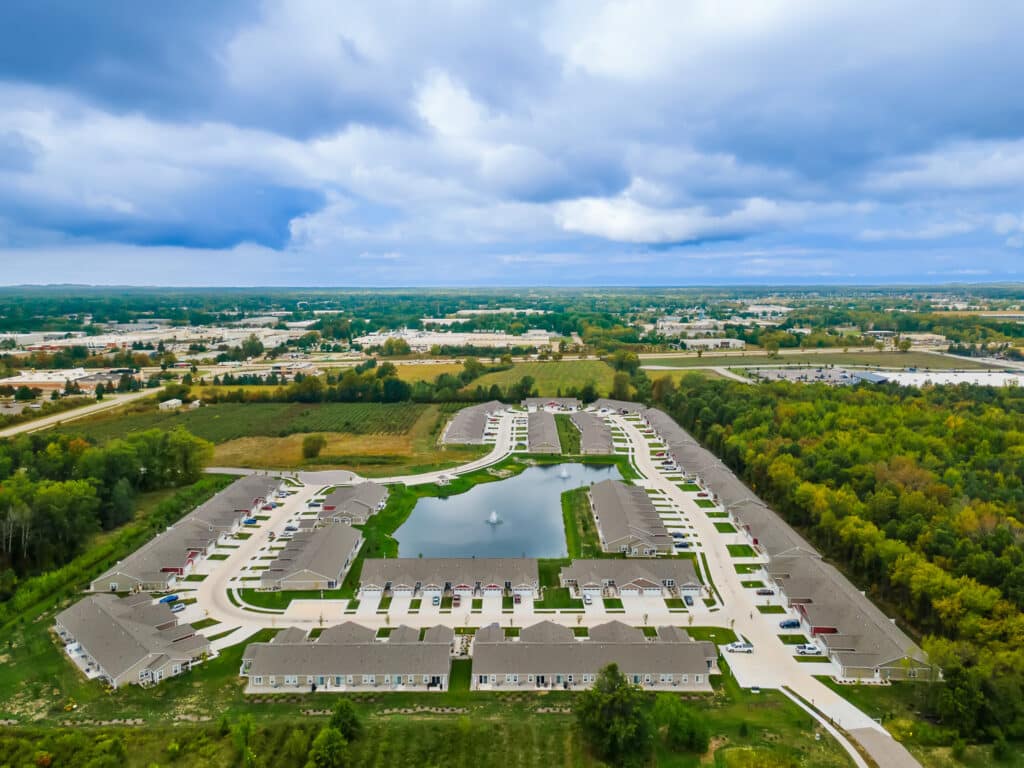 Aerial view of a residential neighborhood with rows of houses surrounding a central pond and fountain, bordered by trees and open land under a cloudy sky.