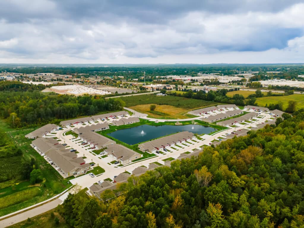 Aerial view of a suburban neighborhood with houses surrounding a small pond, bordered by trees and green spaces under a cloudy sky.