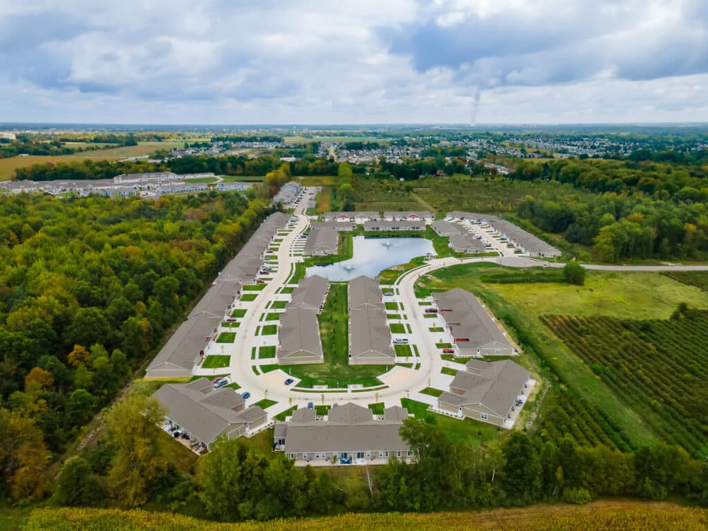 Aerial view of a suburban residential neighborhood with rows of houses, a central pond, surrounding trees, and open fields under a cloudy sky.