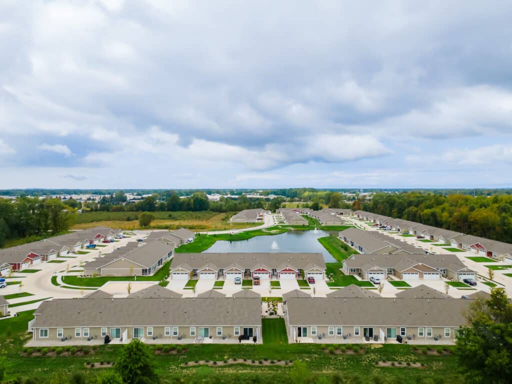 Aerial view of a suburban residential neighborhood with rows of houses, driveways, green spaces, and a central pond under a cloudy sky.