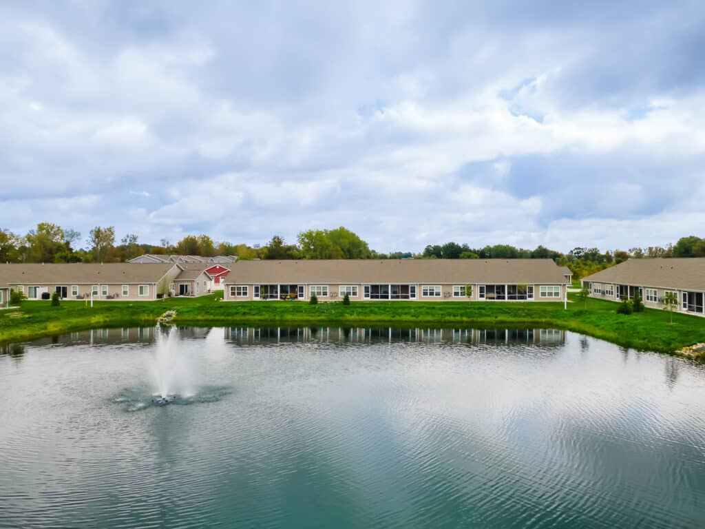 A row of single-story buildings faces a small pond with a fountain in the center, grassy banks, and trees in the background under a partly cloudy sky.