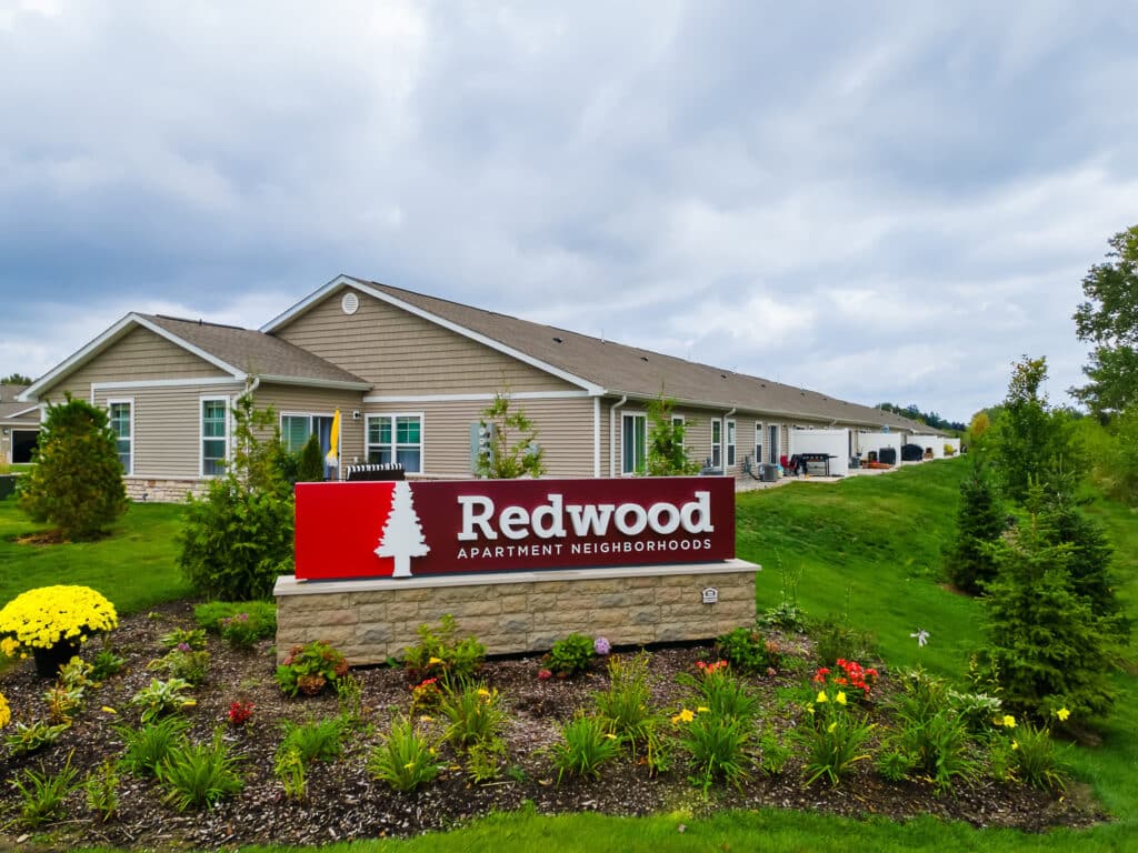 A single-story apartment building with a landscaped garden and a red sign in front reading "Redwood Apartment Neighborhoods.