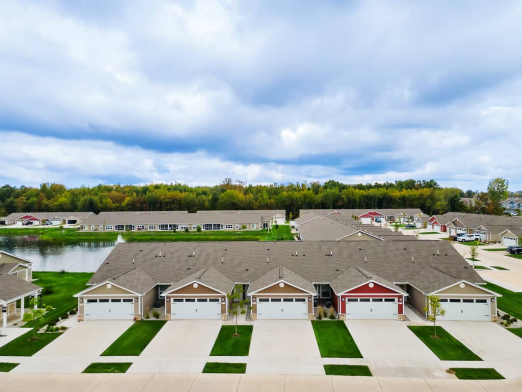 Row of suburban single-story houses with attached garages, green lawns, and a pond in the background under a cloudy sky.