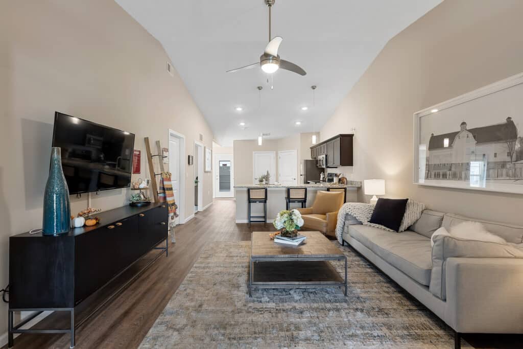 Modern living room and kitchen with neutral tones, a gray couch, wall-mounted TV, coffee table, ceiling fan, and wooden flooring under vaulted ceilings.