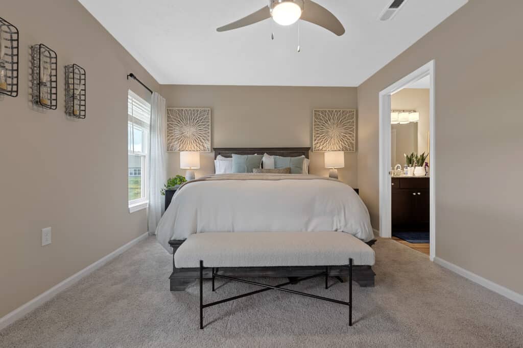 A neatly made bed with a bench at the foot, two nightstands with lamps, wall art, and a ceiling fan in a carpeted bedroom with beige walls.