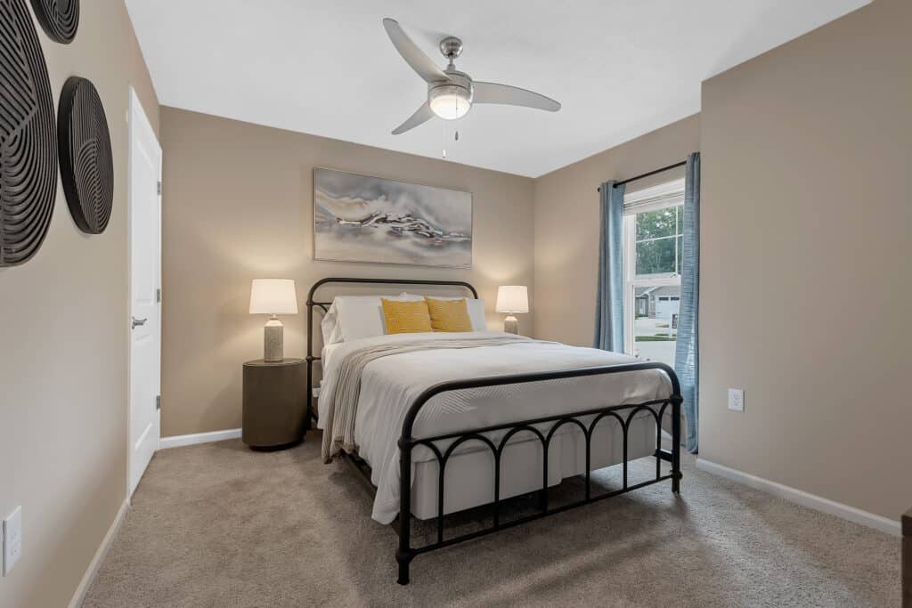 A neatly made bed with yellow accent pillows is centered in a beige bedroom, flanked by two nightstands with lamps, and a ceiling fan and window are visible.