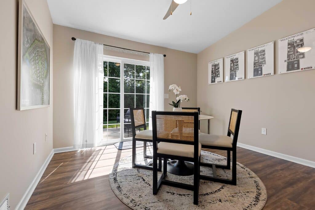 A small dining area with a round table and four chairs on a circular rug, near sliding glass doors overlooking a green outdoor space. Artwork and floor plans hang on the walls.