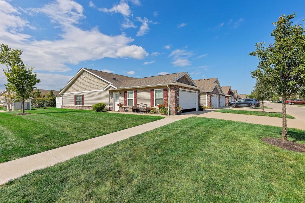 Single-story suburban house with attached garage, front porch, well-kept lawn, trees, and a driveway on a sunny day.