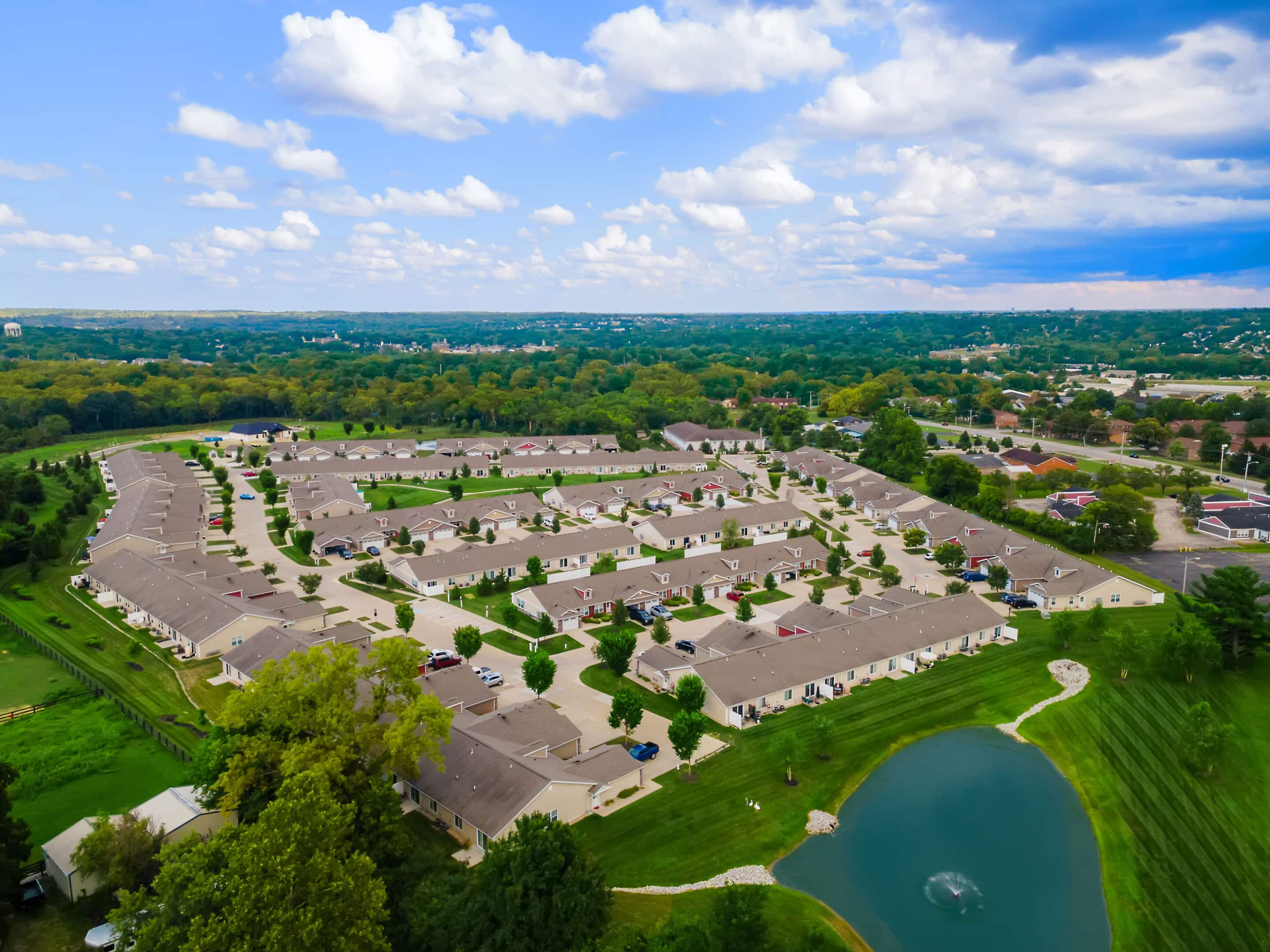 Aerial view of a suburban neighborhood with rows of houses, green lawns, a small pond with a fountain, and trees in the background under a partly cloudy sky.