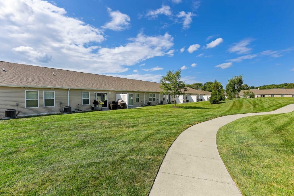A wide sidewalk curves through a neatly mowed lawn behind a row of beige single-story townhomes under a partly cloudy blue sky.
