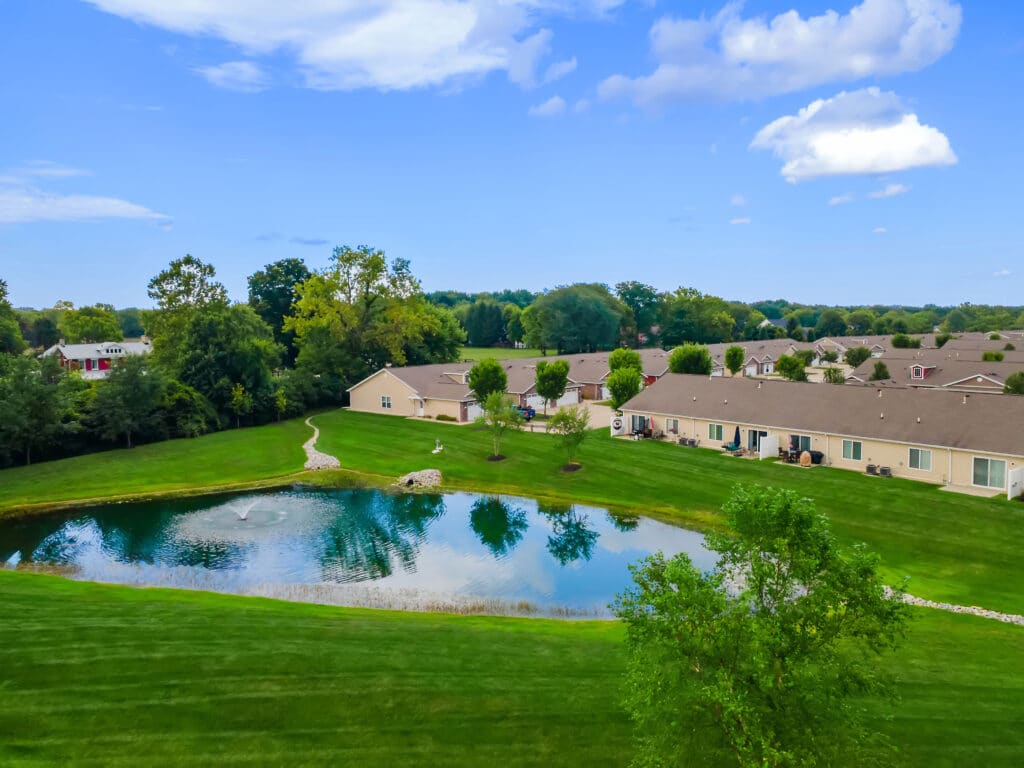 A small pond with a fountain is surrounded by green grass and trees, adjacent to a row of single-story residential buildings under a partly cloudy sky.