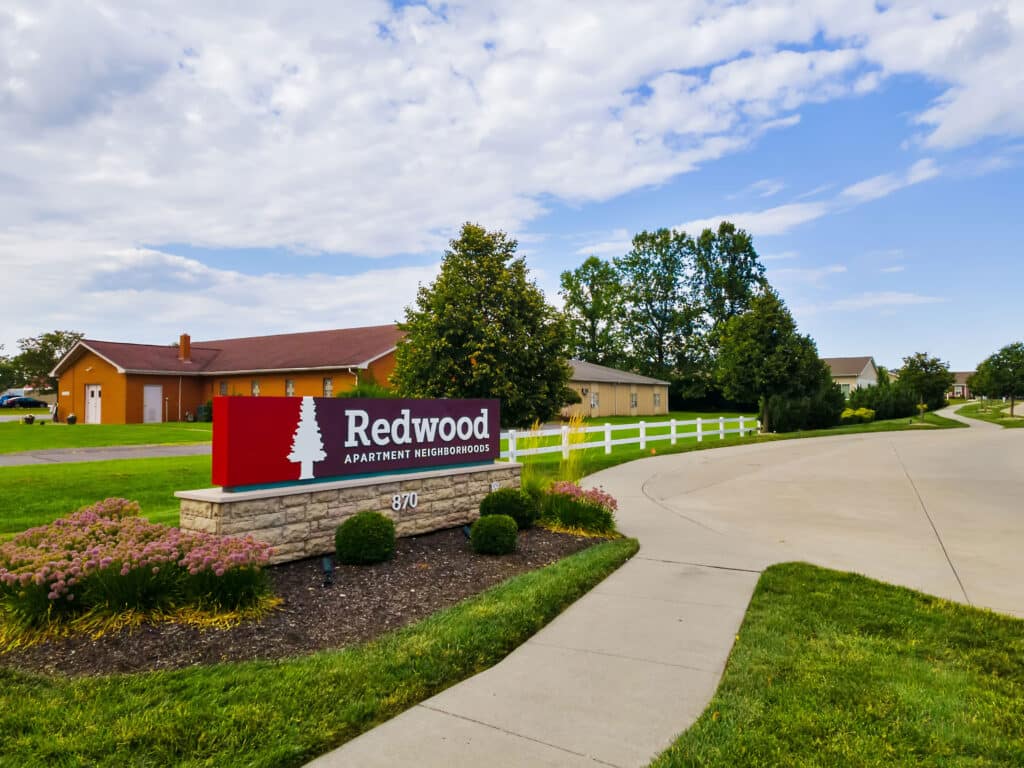 A red and white sign reading "Redwood Apartment Neighborhoods" stands by a driveway, with grass, flowers, and houses in the background under a partly cloudy sky.