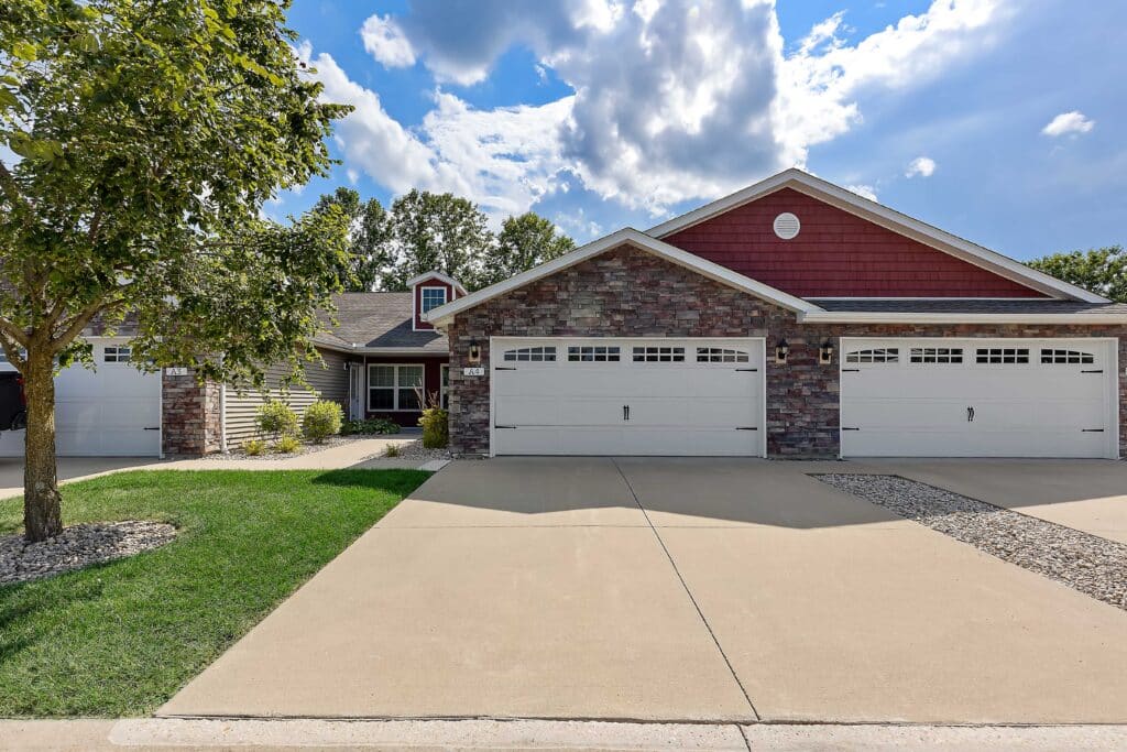 A duplex with two separate garages, stone and red siding, a driveway, a small tree, and landscaped areas, under a partly cloudy sky.