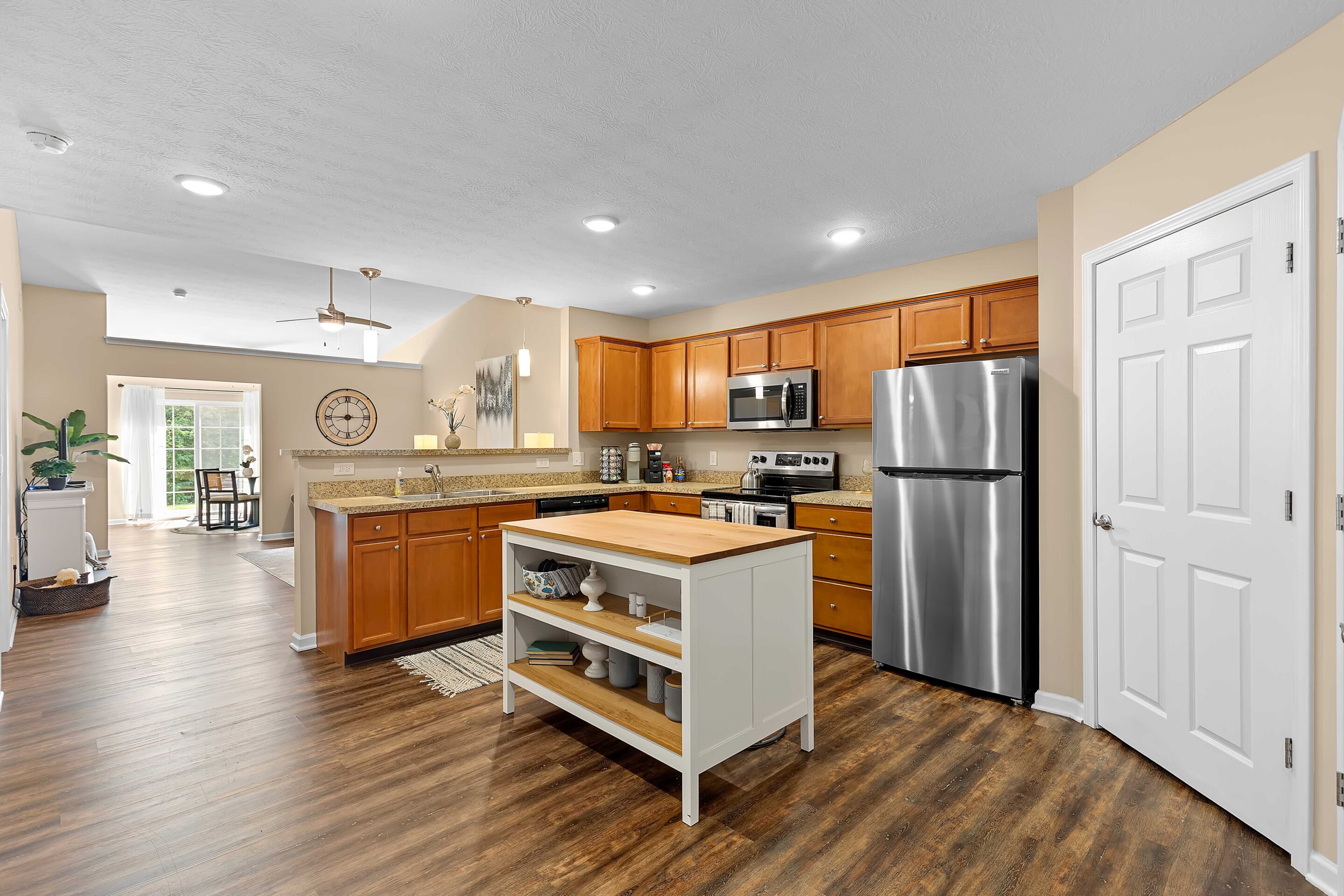 Modern kitchen with stainless steel appliances, wooden cabinets, a central island with shelves, and wood flooring; a dining area is visible in the background.