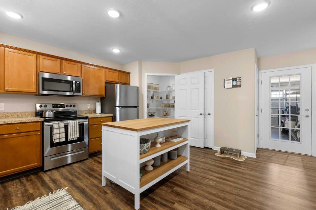 A modern kitchen with wood cabinets, stainless steel appliances, a white island, a pantry with open shelves, and a door leading to a garage.