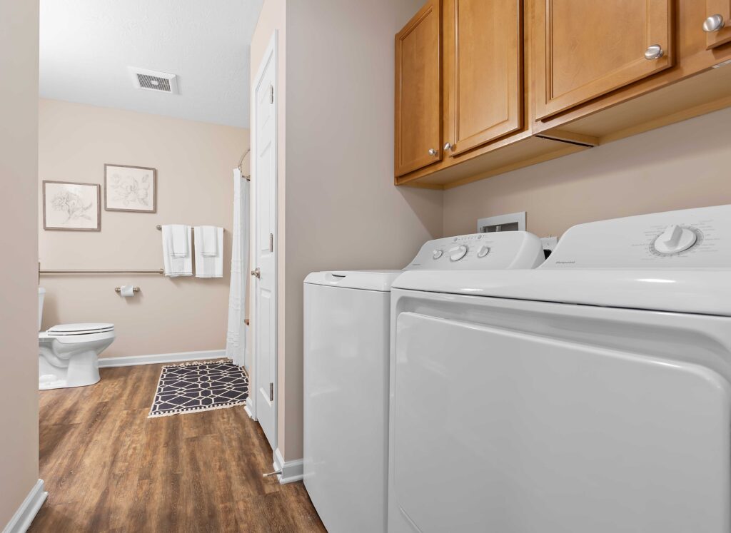 A laundry area with a washer and dryer next to a bathroom featuring a toilet, towel rack, wall art, and wood-style flooring.