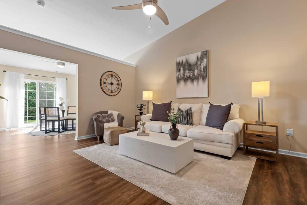 A modern living room with a beige sofa, armchair, coffee table, and two lamps. Large window in the adjacent dining area lets in natural light. Walls and decor are neutral-toned.