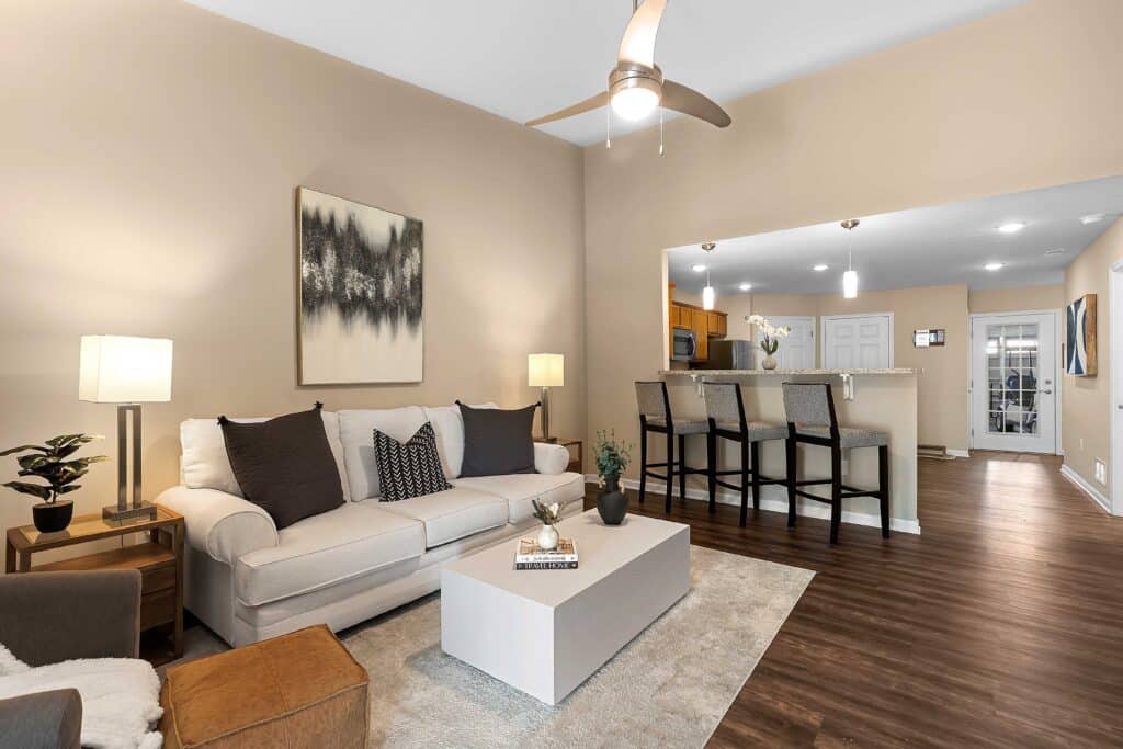 Modern open-concept living room and kitchen with neutral tones, a white sofa, bar stools at the counter, ceiling fan, and contemporary decor.