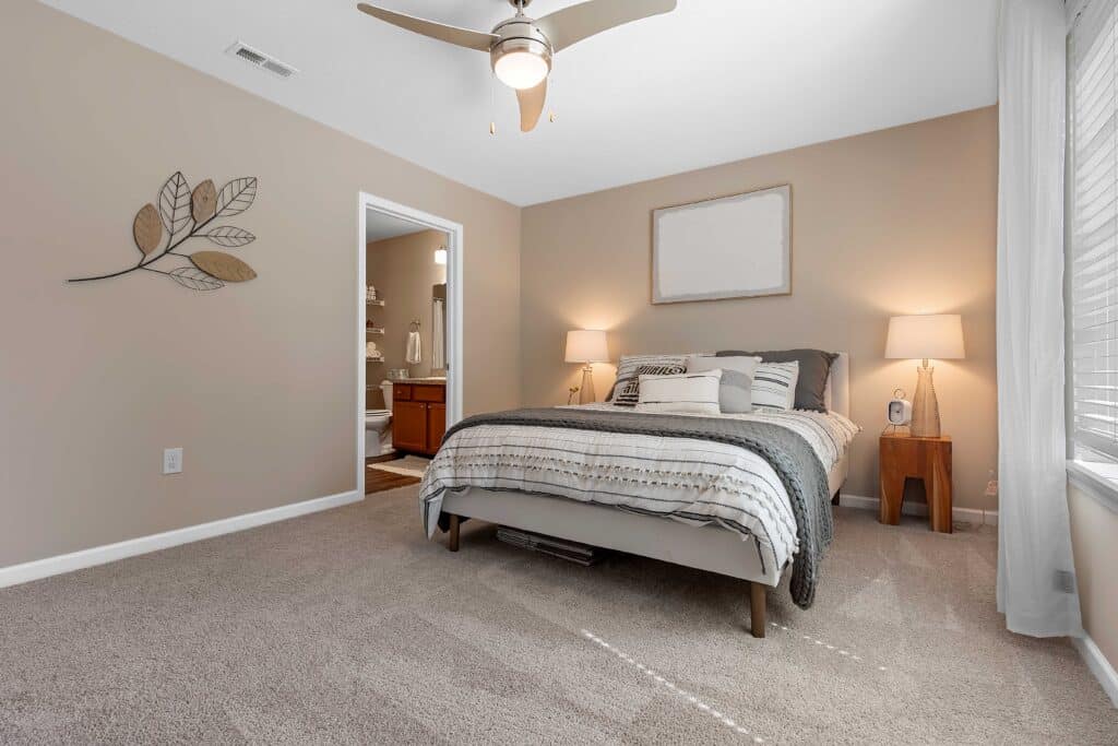 A neatly made bed with neutral bedding in a carpeted bedroom, two bedside tables with lamps, wall art, and an open doorway leading to a bathroom.