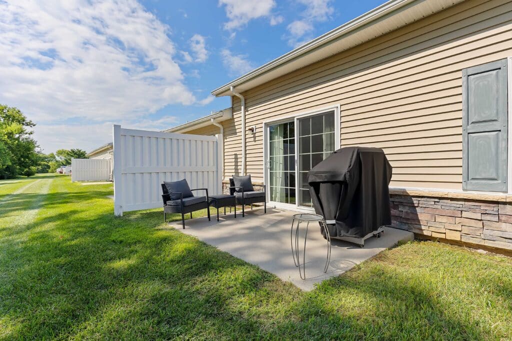 Small backyard patio with two chairs, a round table, and a covered grill next to a sliding glass door; white privacy fence and grass lawn surrounding the area.