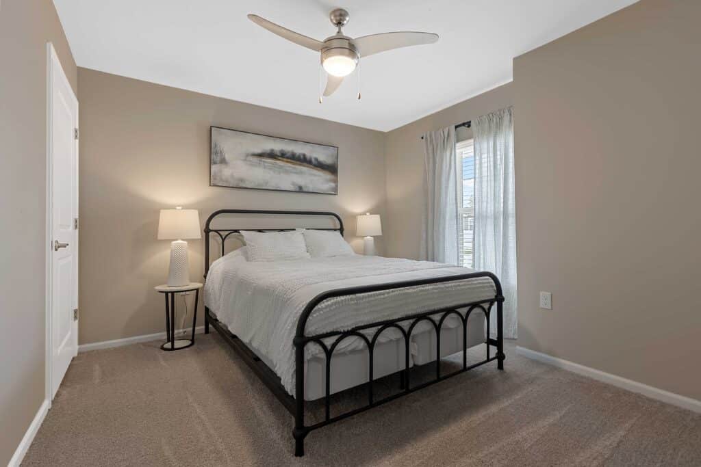 A bedroom with a black metal bed frame, white bedding, two lamps on side tables, beige walls, carpeted floor, ceiling fan, and a window with sheer curtains.