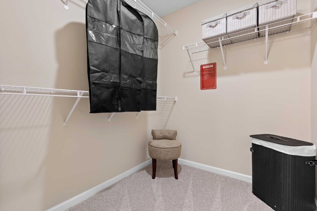 A walk-in closet with wire shelving, a black garment bag, two storage baskets, a cushioned chair, a black laundry hamper, and beige carpet flooring.