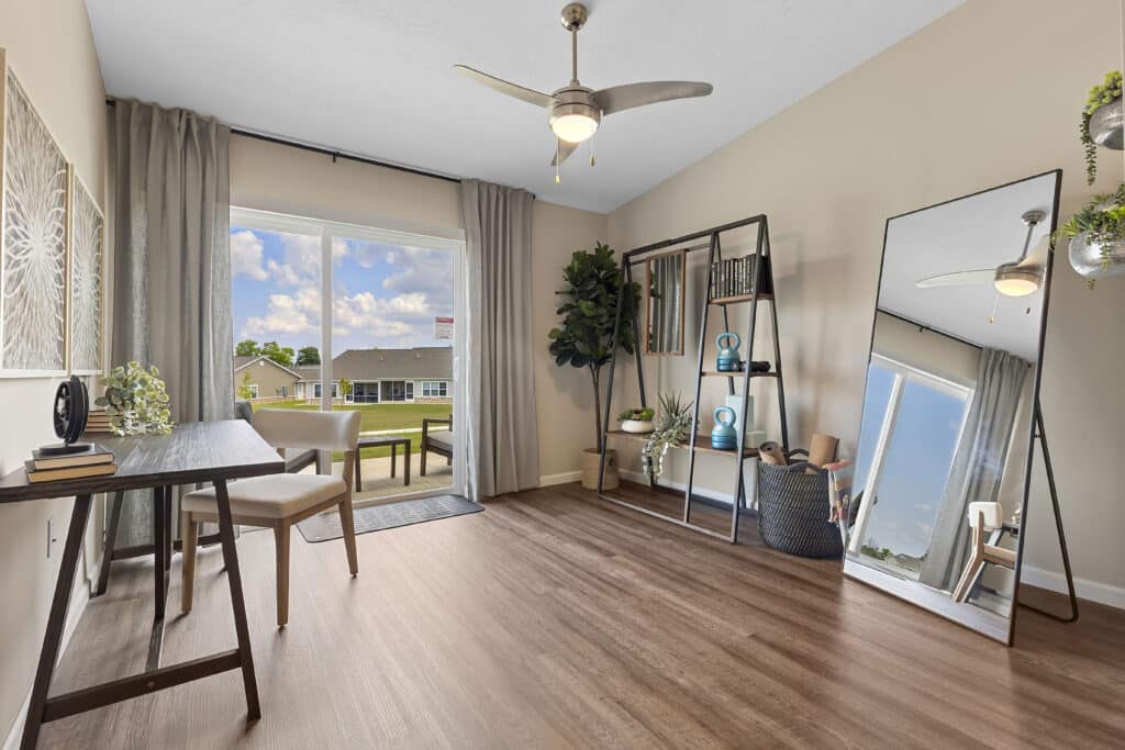 A modern home office with a desk, chair, bookshelves, large mirror, indoor plants, and floor-to-ceiling window overlooking a suburban neighborhood.