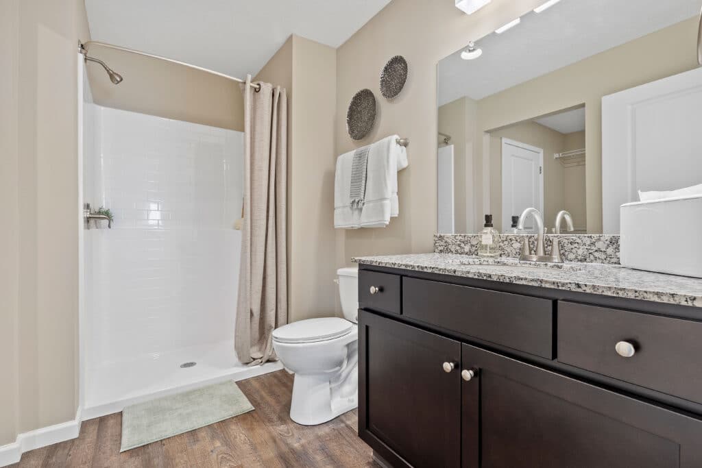 Modern bathroom with a walk-in shower, granite countertop vanity, dark wood cabinets, toilet, and towels neatly arranged on the wall.