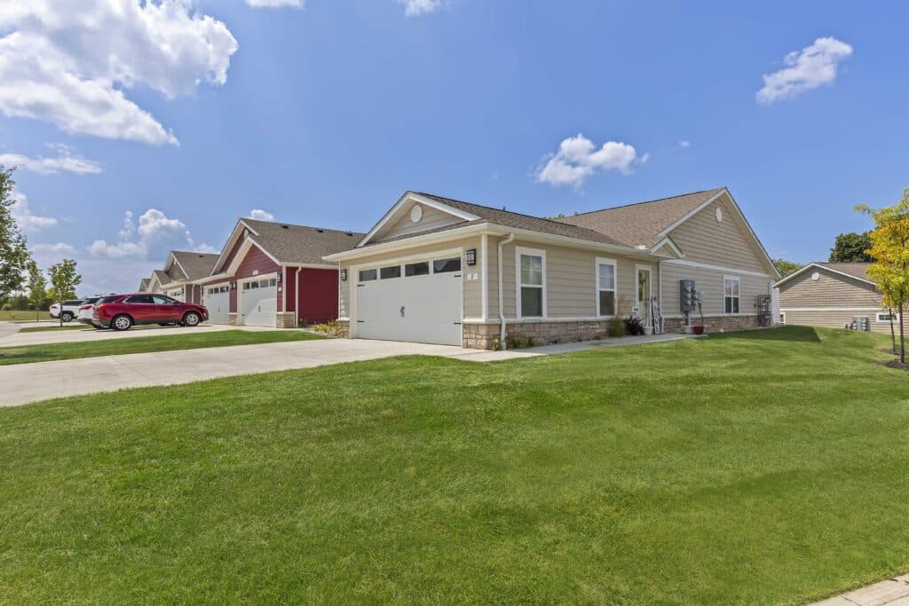 Single-story suburban house with an attached two-car garage, beige siding, and a well-maintained lawn under a blue sky with scattered clouds.