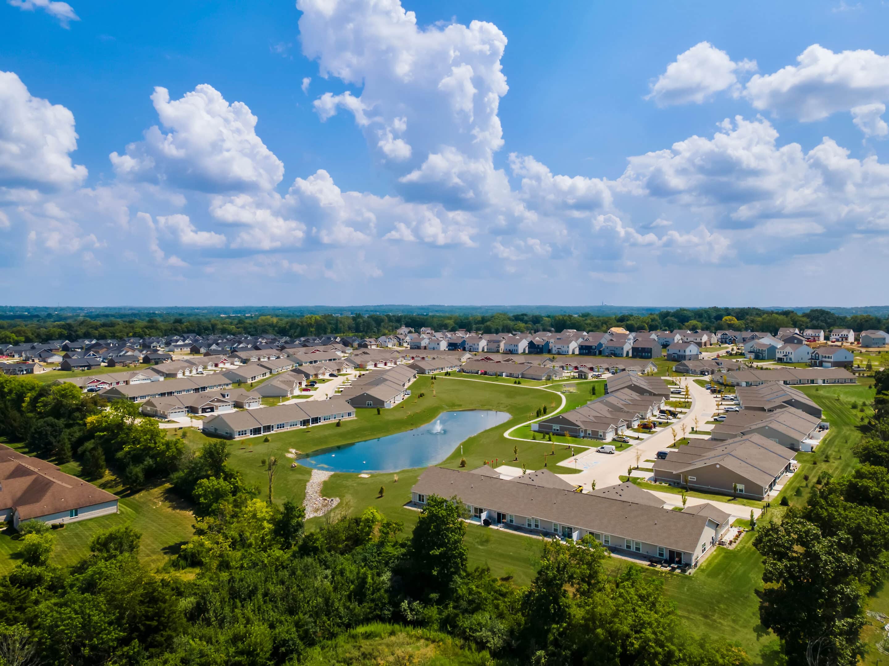 Aerial view of a suburban neighborhood with rows of houses, a small pond, green lawns, trees, and a partly cloudy sky.