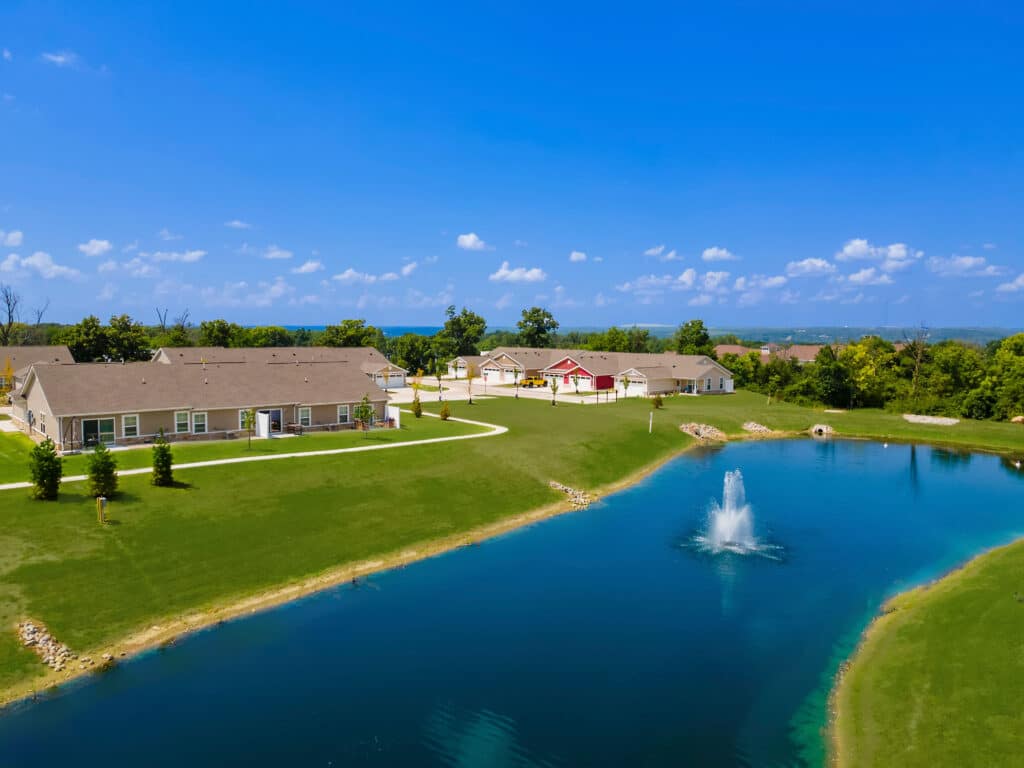 A small pond with a fountain is surrounded by green grass and single-story homes under a clear blue sky.