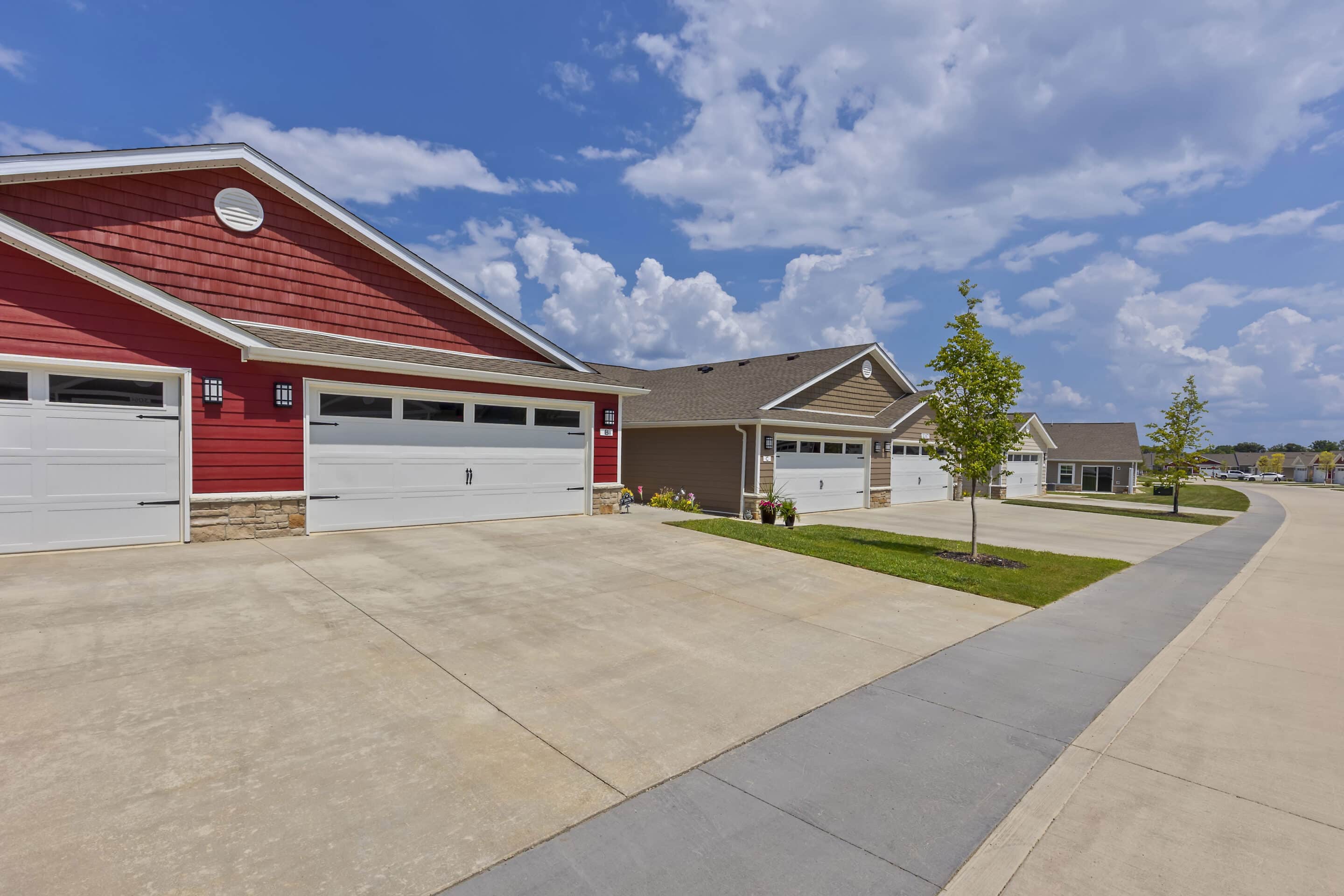 Row of modern suburban houses with attached garages, concrete driveways, and small newly-planted trees along a clean residential street under a partly cloudy sky.