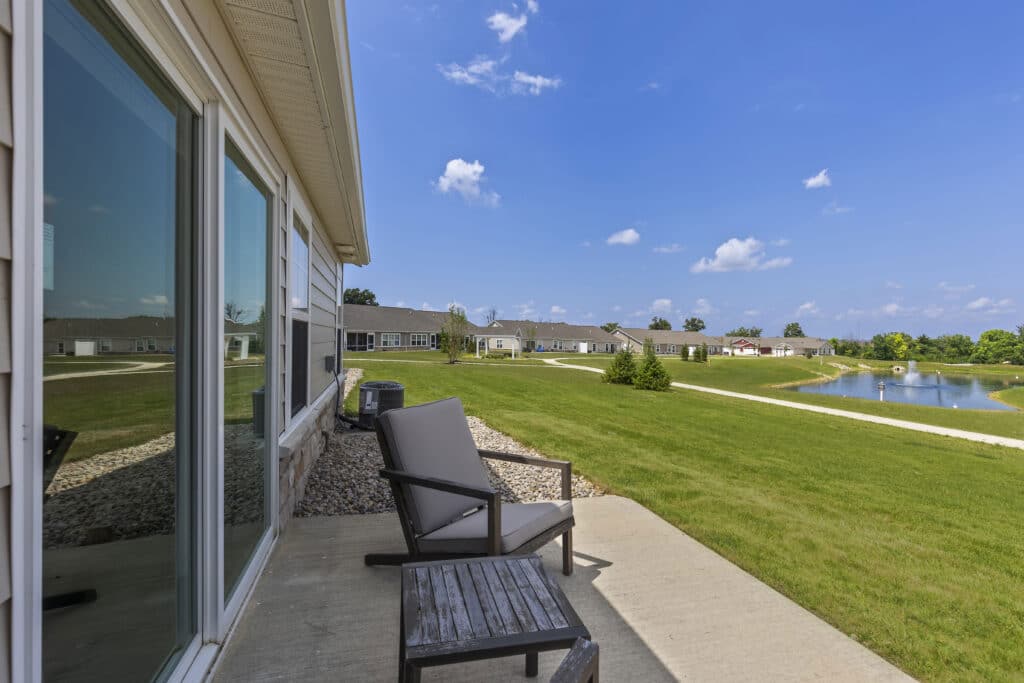 A patio with two chairs overlooks a grassy lawn, pond, and a row of single-story homes under a blue sky with scattered clouds.