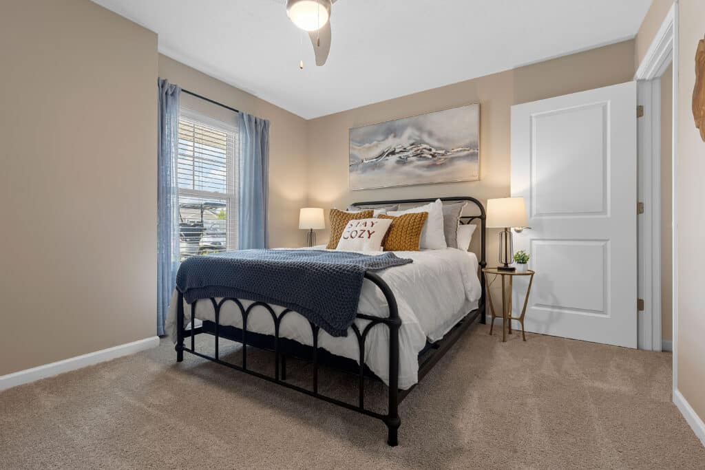A neatly made bed with pillows, a blanket, and "STAY COZY" cushion is in a carpeted bedroom with beige walls, lamps, a window with curtains, and a painting above the bed.