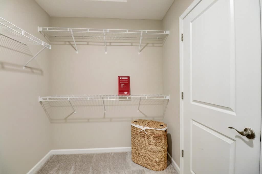 Empty walk-in closet with white wire shelves, beige carpet, a closed white door, and a wicker laundry basket on the floor.