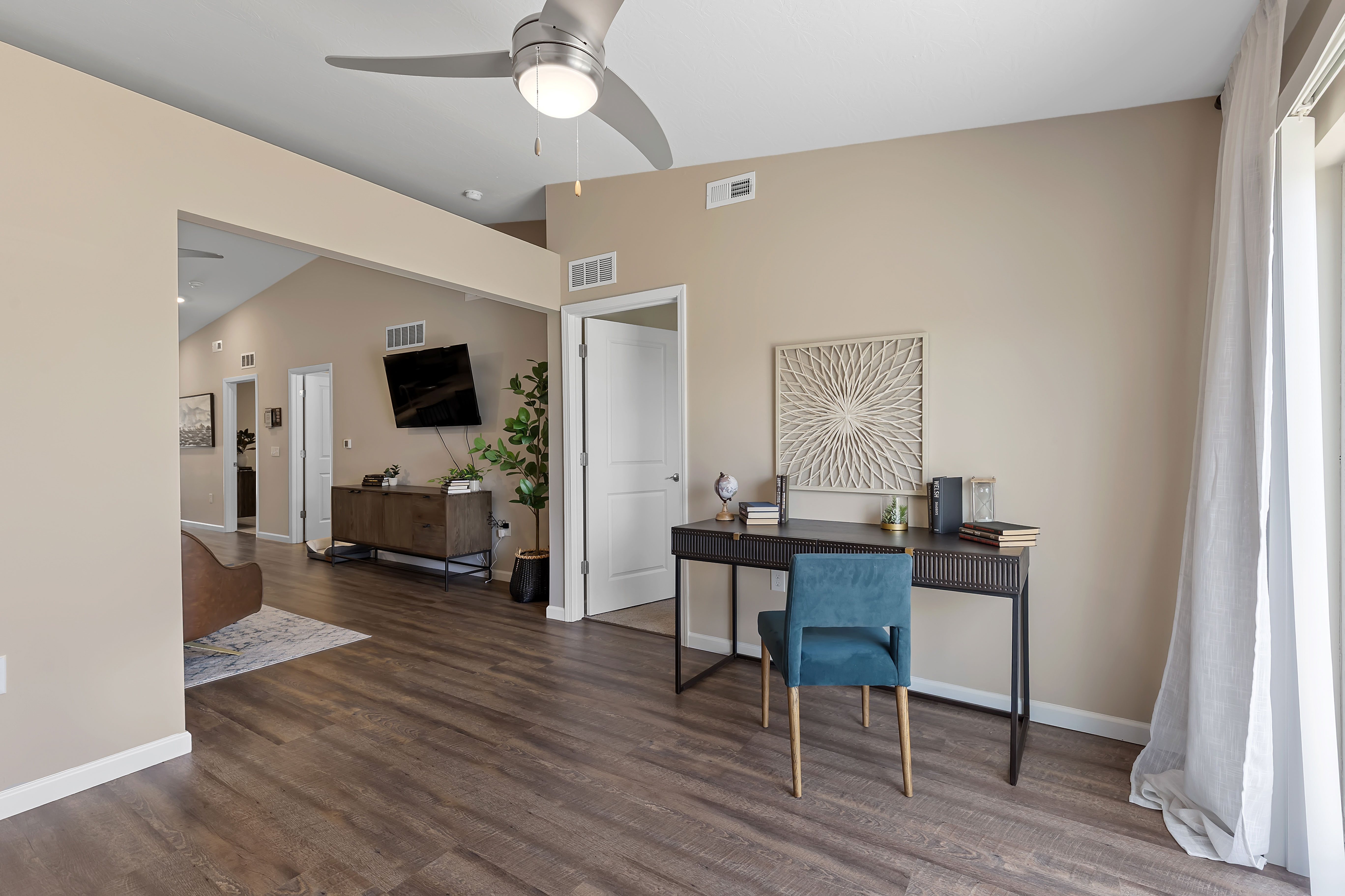 A modern home office space with a black desk, blue chair, books, and decor, adjacent to a living area with wood flooring and beige walls.