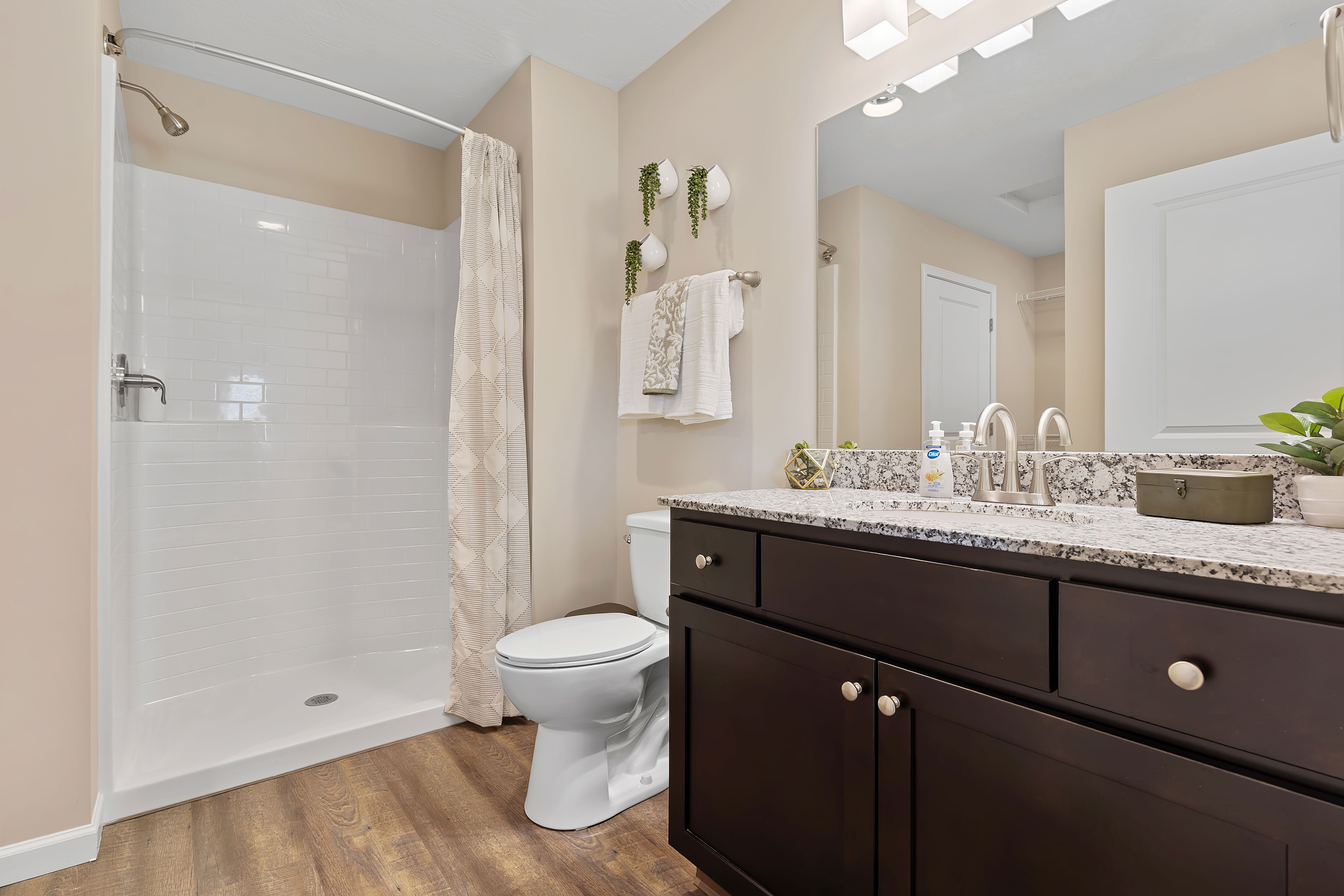 A modern bathroom with a walk-in shower, toilet, dark vanity with granite countertop, large mirror, and wood-style flooring.